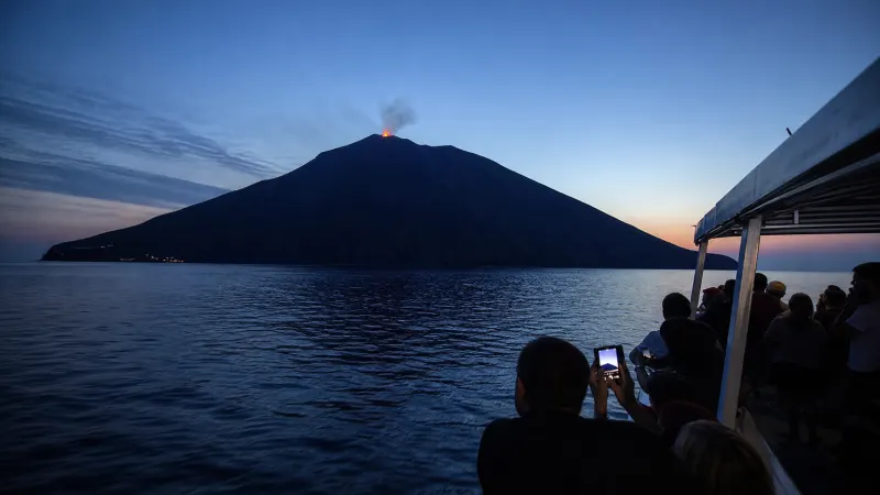 Stromboli di notte da Tropea: barca in navigazione di fronte al vulcano con il bagliore di un'esplosione al crepuscolo