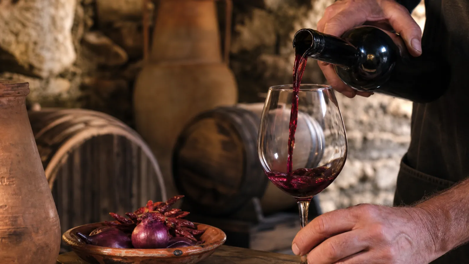 Mani di un vignaiolo che versa vino rosso in una cantina di pietra con botti, accanto a cipolle rosse e peperoncini calabresi